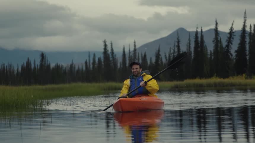 Man in orange kayak paddling on calm water surrounded by trees and mountains under cloudy sky