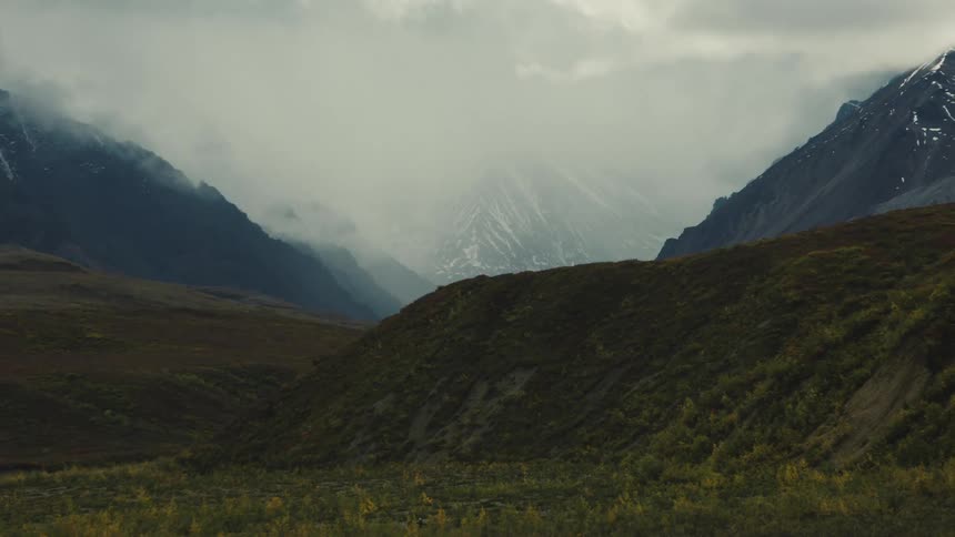Lush green hill in foreground with valley flanked by steep mountains and snowy peaks in the background under cloudy sky