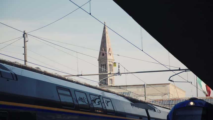 From a train station platform a historic church steeple rises above the city framed by the silhouettes of modern trains and a web of overhead power lines