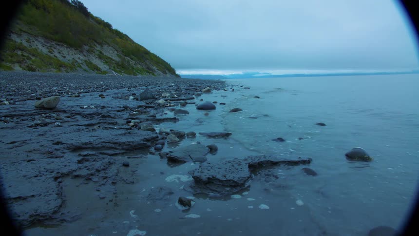 Rocky shoreline with scattered stones green cliff on the left and calm water under cloudy sky