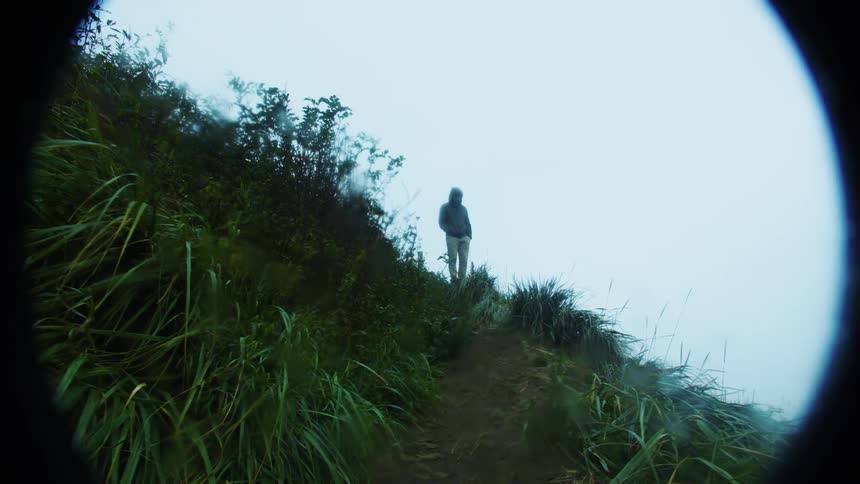A silhouetted human figure stands on a narrow dirt path on a steep green hillside as thick mist obscures the background all framed by a circular visual border