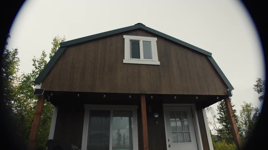 Wooden cabin with white windows surrounded by vegetation under a cloudy sky