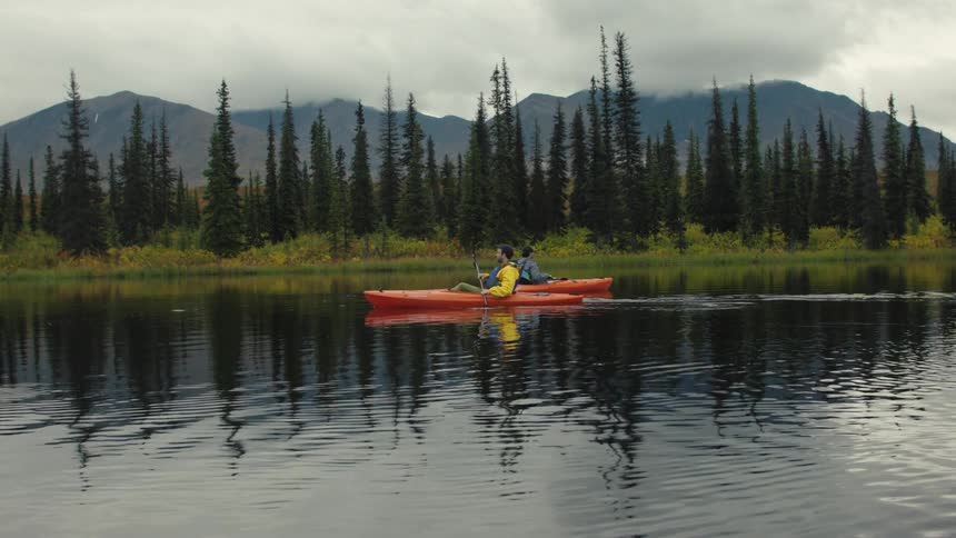 Two people in an orange tandem kayak paddle slowly across a calm lake surrounded by a dark pine forest and mountains under an overcast sky the waters surface reflects the trees and the slight ripples from the boat006