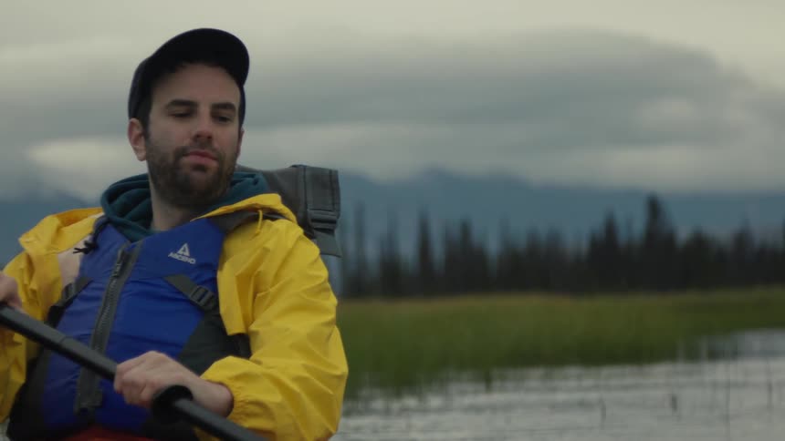 Man wearing a baseball cap paddling on a kayak close to the shore with a forest and hills on the background outdoors during day time