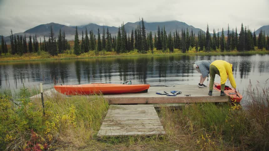 Two men carry a kayak to the river from the dock at midday setting it in the water as one prepares to board wearing a life jacket