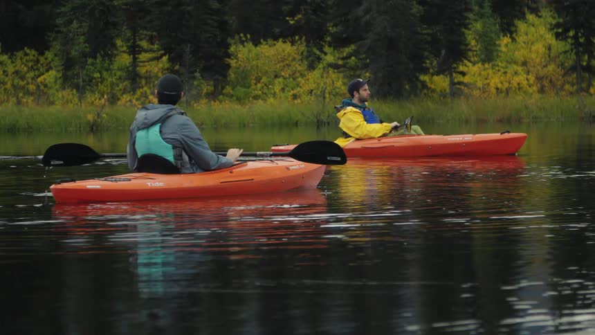 Two people in orange kayaks paddling on calm water surrounded by trees under cloudy sky