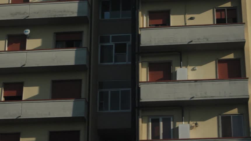 The video shows a quick closeup lateral dolly shot of the balconies and windows of an aging multistory apartment building under a clear blue sky the camera moves rapidly past the facade suggesting a view from a passing vehicle