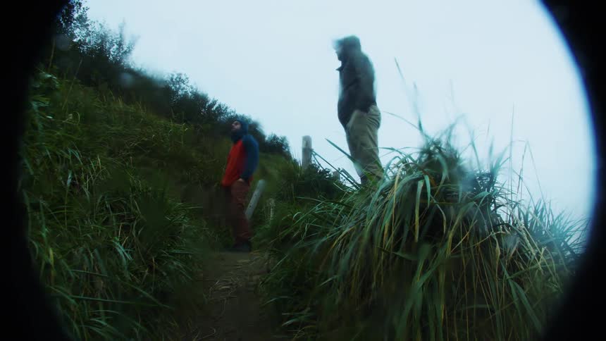 Two people dressed in outdoor gear stand on a narrow overgrown mountain trail that cuts across a grassy slope against an intensely overcast or foggy sky the footage is framed with a distinct binocularlike vignette suggesting they are being observed