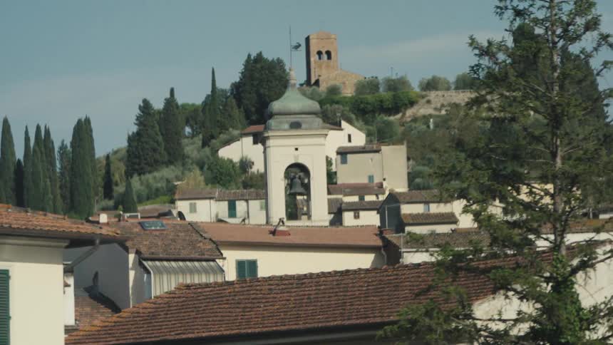 A slow moving video from a car shows the redtiled rooftops of a small italian town with a white bell tower topped by a dome and a distant medieval brick tower on a treecovered hill framed by blurred foliage in the foreground
