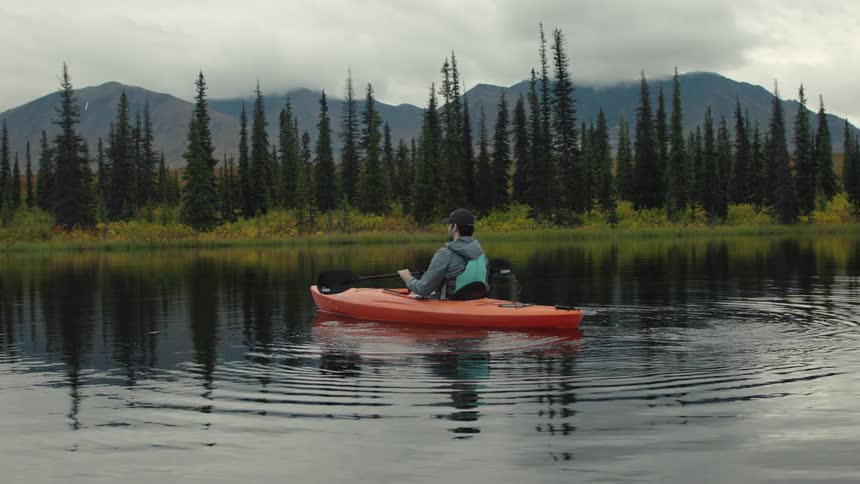 Person paddles a bright orange kayak on a calm lake surrounded by pine trees and mountains