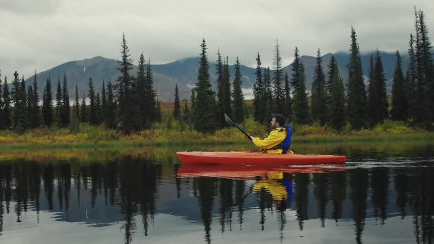 Person in a yellow jacket paddles a bright orange kayak on a lake surrounded by pine trees and mountains