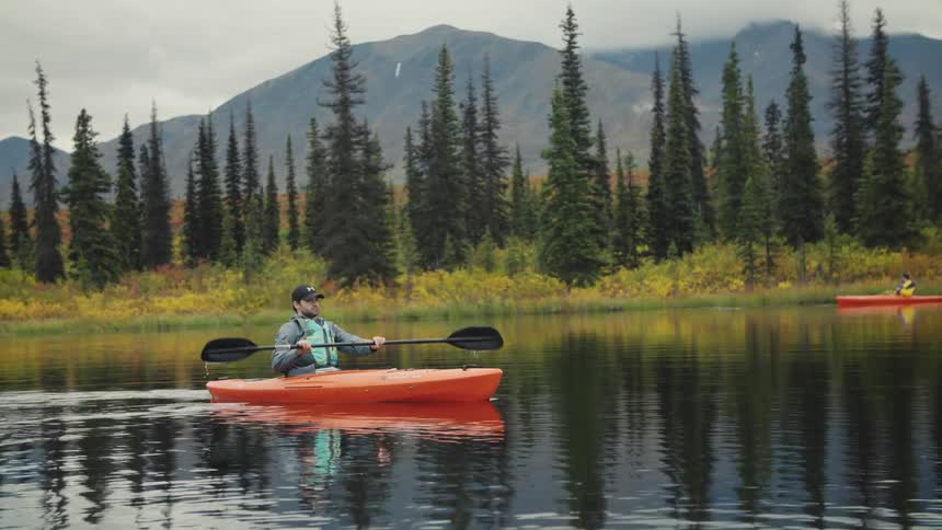Man in a blue vest paddles an orange kayak on a calm lake surrounded by pine trees and mountains