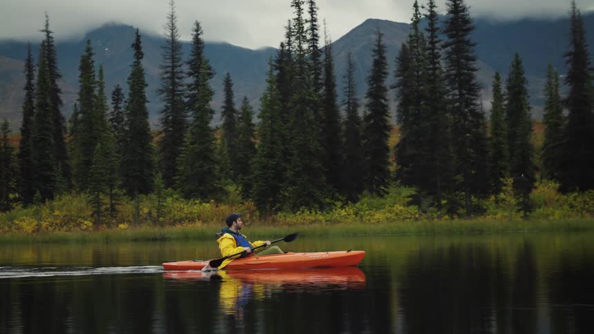 Person in orange kayak on calm lake surrounded by trees and mountains with clouds in the background