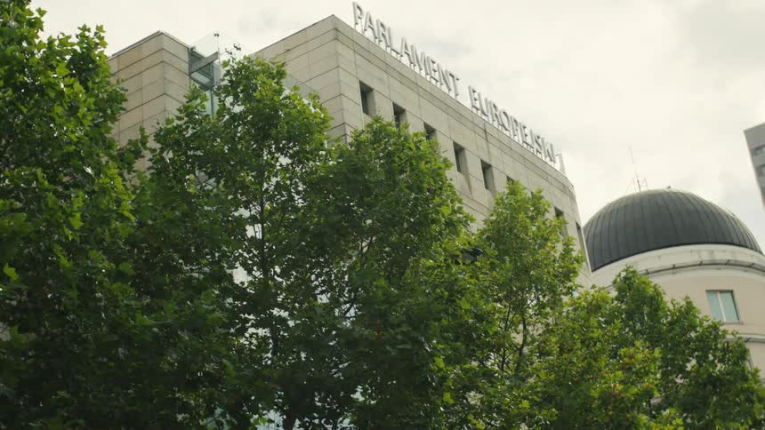 The video shows a lowangle view of a modern building with parlament europejski visible in large letters at the top mostly framed by a row of bright green trees the camera slowly pans right revealing a white building with a dark dome next to it