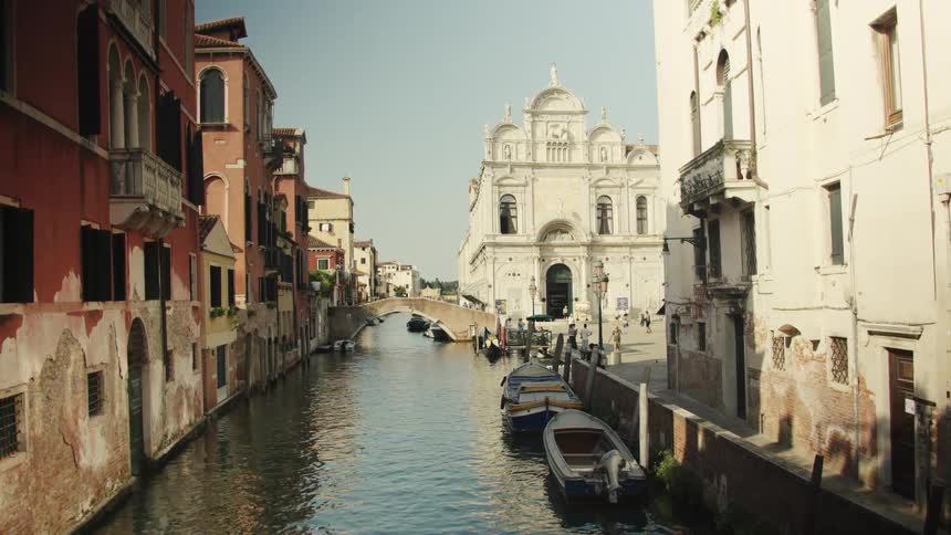 A quiet canal lined with weathered stucco buildings and moored boats leads to a grand white church and small stone footbridge