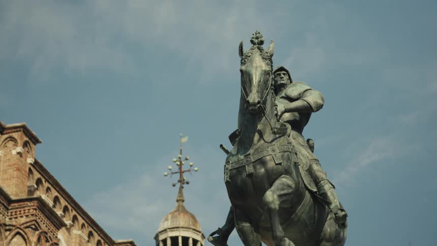 The video is a lowangle static shot of a large bronze equestrian statue of a man in armor likely a condottiero or military leaderbehind the statue against a mostly clear sky a building facade with crenellations and a small dome or cupola