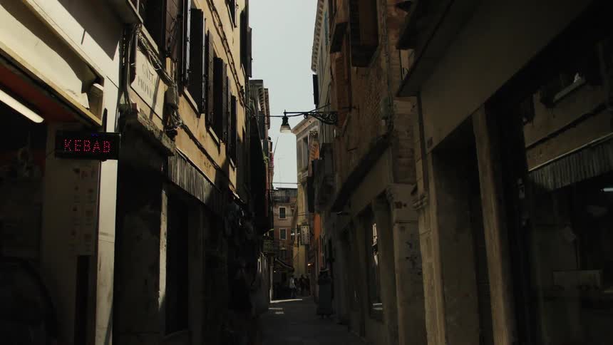 A lowangle video captures the dramatic vertical perspective of a narrow shadowed alleyway between tall aging buildings in a european city leading to a brighter distance where pedestrians are faintly visible beneath an overhead street lamp