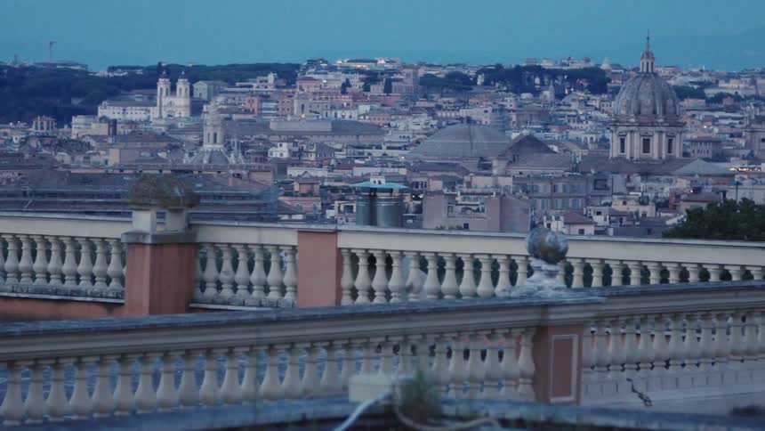 The video is a sweeping panoramic view of a large european city skyline likely rome taken from an elevated vantage point at dusk showing a densely packed urban area with numerous historic domes and architectural landmarksthe foreground features a de