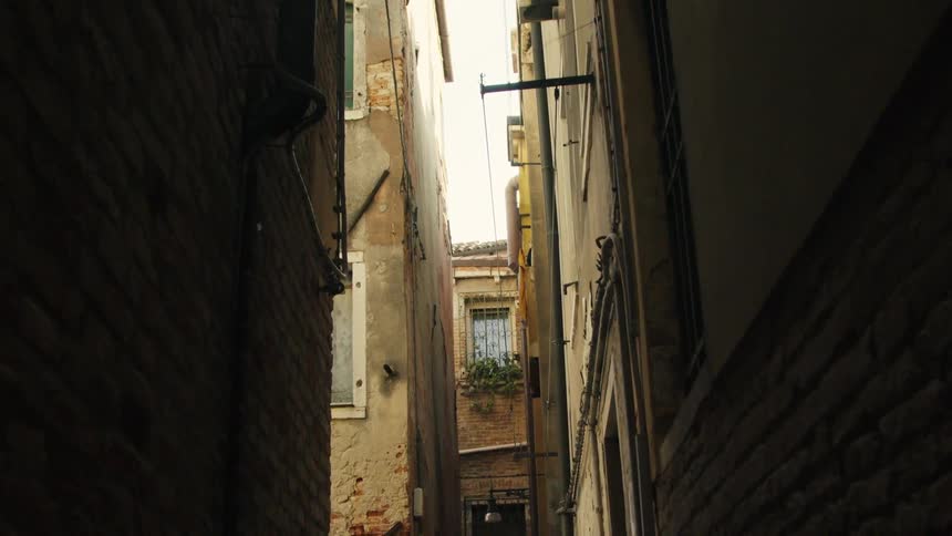 The camera slowly pans down a remarkably narrow dark alleyway between tall aged buildings likely in venice italy the walls feature weathered brick and stucco with a single visible window holding a small plant box