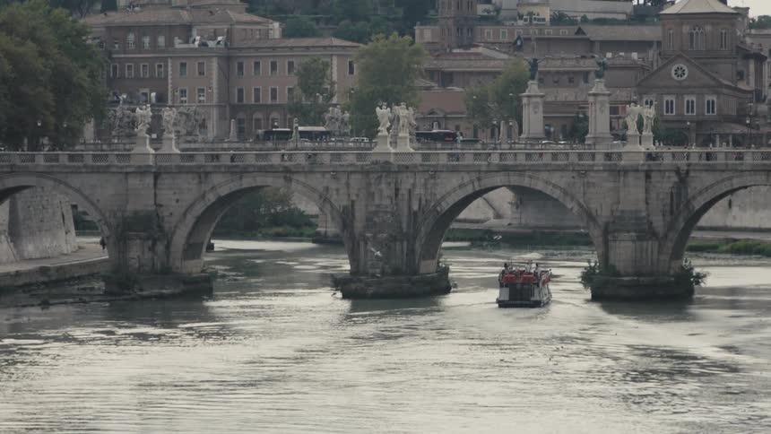 A stone bridge lined with statues spans a calm river while a small tour boat passes beneath backed by historic buildings