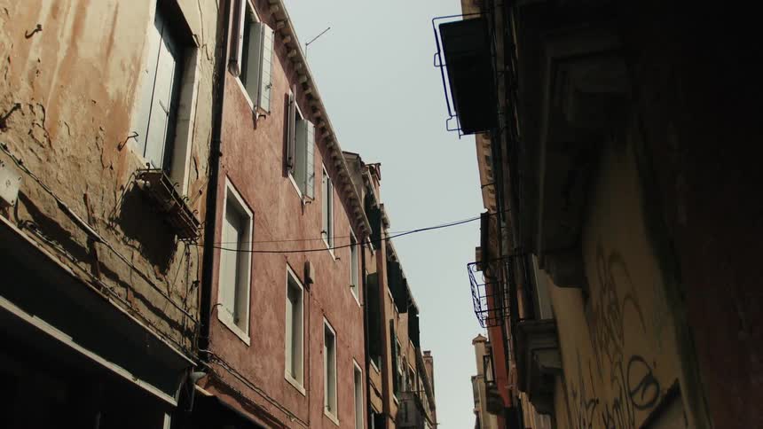 The video is an extreme lowangle shot looking up at the narrow gap between two rows of tall historic buildings in an italian city likely a venetian callethe buildings on the left have faded reddishorange plaster walls while the darker buildings