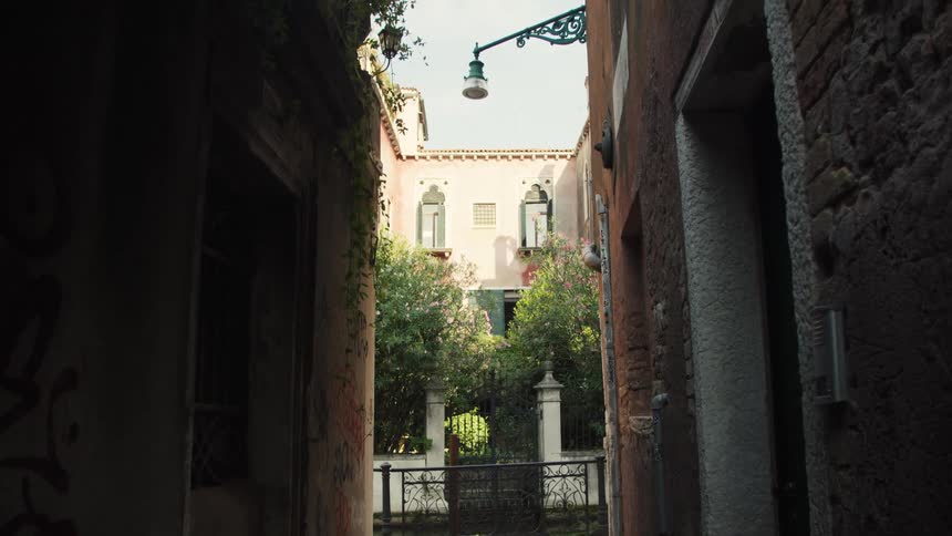 The video shows a view looking out from a dark narrow alleyway framed by old stone or brick walls on both sidesthe alley opens up to a bright small courtyard area with green trees and a black iron fence or gate beyond which stands a lightcolored