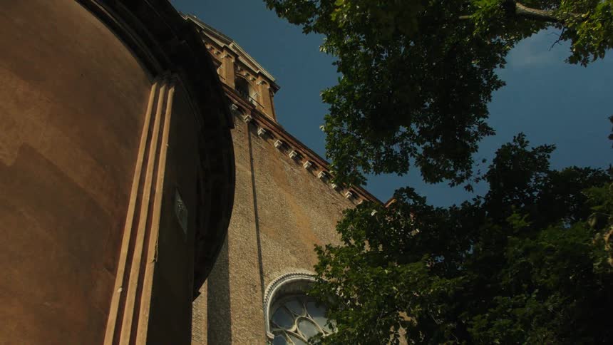 Upward view of a warmtoned brick church wall with a tall gothic arched window side buttress and leafy tree catching lateafternoon light