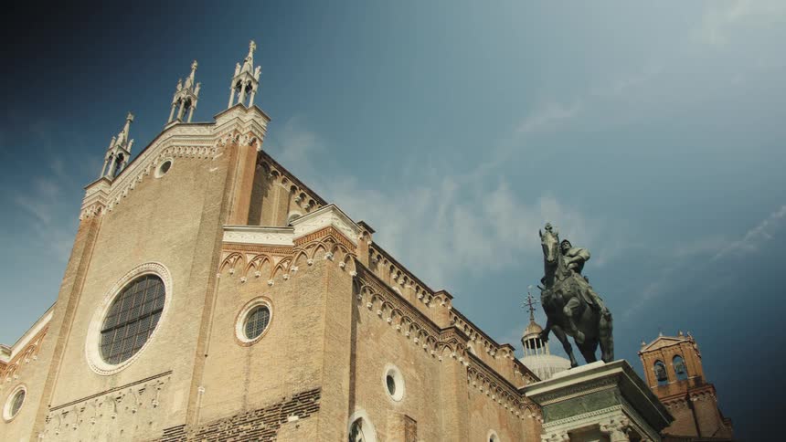 A lowangle view video slowly pans across the gothic brick facade and ornate pinnacles of a large church and a prominent bronze equestrian statue on a tall pedestal in the foreground against a blue and cloudy sky