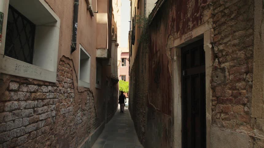 A narrow sunlit alley of worn brick and stucco leads toward a bright courtyard where a lone pedestrian walks