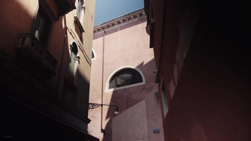 A narrow sunlit alley between pink stucco walls leads toward a faade with a roundarched window under a clear sky