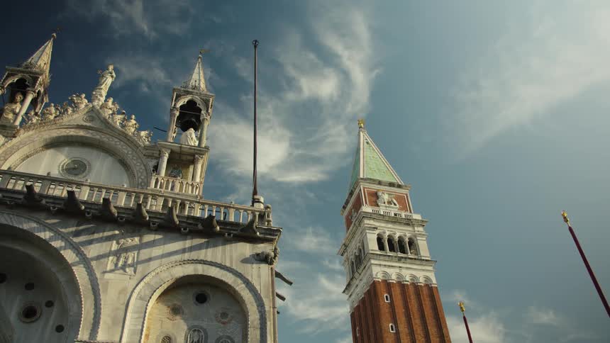 An upward view of ornate white basilica details beside a tall brick bell tower set against a blue sky with wispy clouds