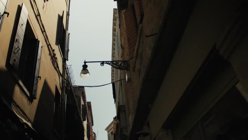 A narrow alley winds through a cityscape of tightly packed residential buildings bathed in the midday sun