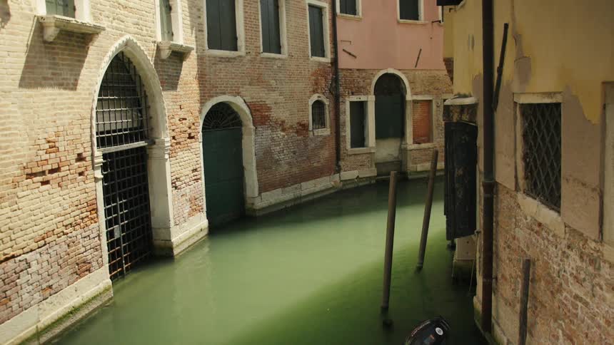 A narrow oldcity canal with greenish water runs between weathered brick buildings arched doorways and leaning wooden mooring posts