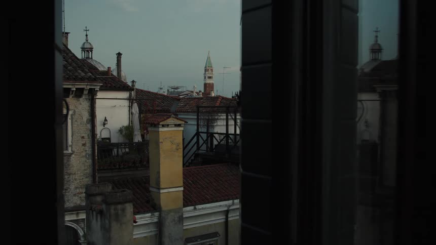 Through a dark window frame terracotta rooftops chimneys and a distant bell tower rise against the evening sky