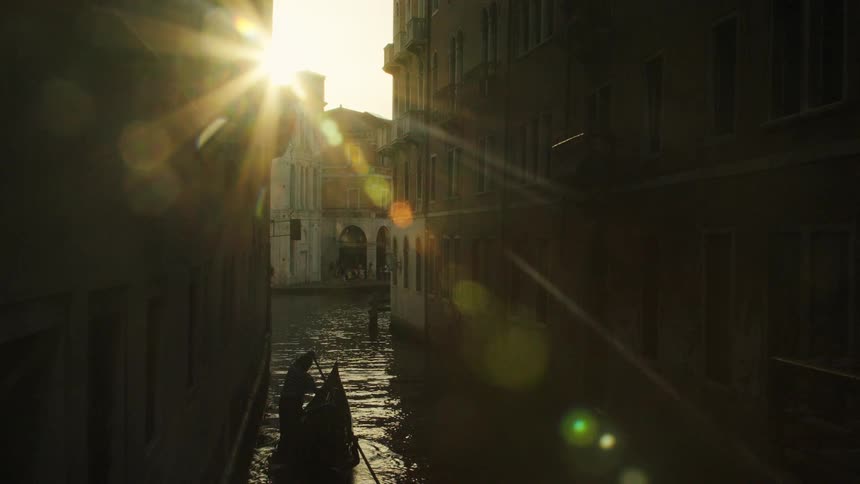 A dark silhouetted gondola with a gondolier rowing moves down a narrow venice canal between tall buildings with a dramatic sunburst and lens flare brightly shining into the camera from the light at the end of the channel