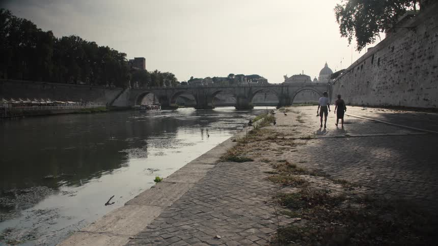 Man and woman walk along riverside path by the tiber with bridges and dome in the background