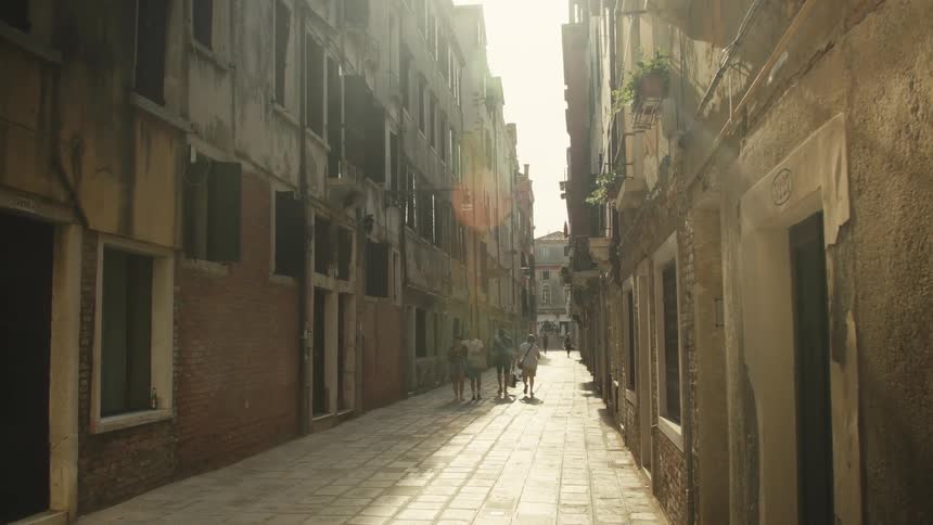 A young man and woman walk away from the camera down a narrow sunlit stone alleyway in venice casting long shadows between tall aging buildings with closed shutters on a bright day as the video slowly tilts up toward the bright sky