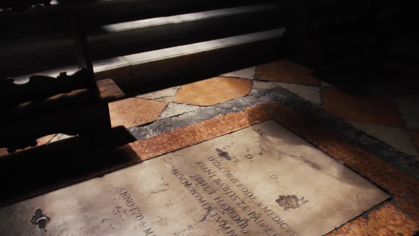A shaft of light falls across a worn stone floor inside a church illuminating a large engraved slab with latin text while nearby pews cast long cool shadows