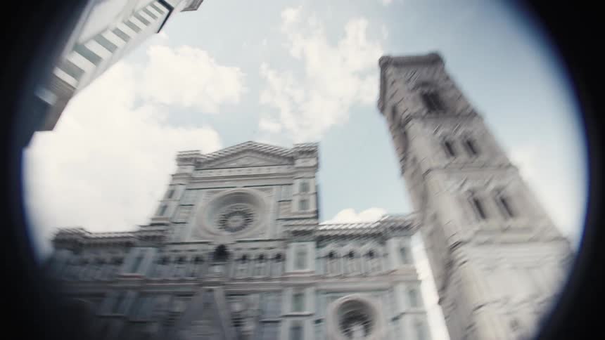 A lowangle fisheye lens video slowly reveals the richly detailed white pink and green marble facade of the duomo cathedral and its adjacent bell tower in florence framed by surrounding buildings against a bright cloudy sky