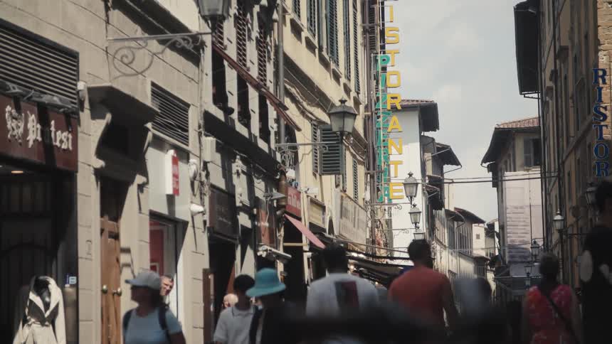 A lowangle video pans slowly across a crowded narrow city street lined with tall aged buildings featuring shops and closed shutters focusing on a partially obscured vertical yellow sign and indistinct pedestrians walking in the foreground