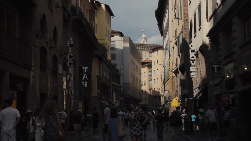 A bustling crowd of pedestrians meanders through a narrow shadowy street flanked by historic european buildings
