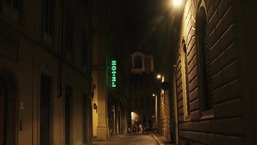 A video focuses on a dark narrow cobblestone alleyway at night lined with tall buildings illuminated by a single bright street lamp and a vertical green neon hotel sign as two indistinct people walk into the distant shadows