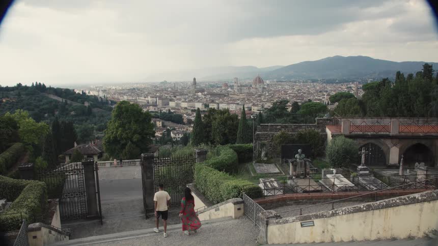 Panoramic overlook of florence with the duomos red dome and palazzo vecchio on the skyline seen from terraced gardens and stone graves as two people descend the steps under a hazy sky