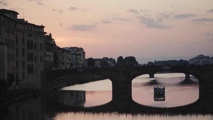 Boat with passengers sails under arched bridge reflected in the river at sunset with historic buildings on the banks
