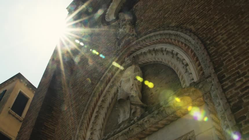 The video is an extreme lowangle shot of the brick facade of an old european church or religious building focusing on an elaborately carved stone archway entrancethe sunlight bursts brightly around the corner of the building creating a drama