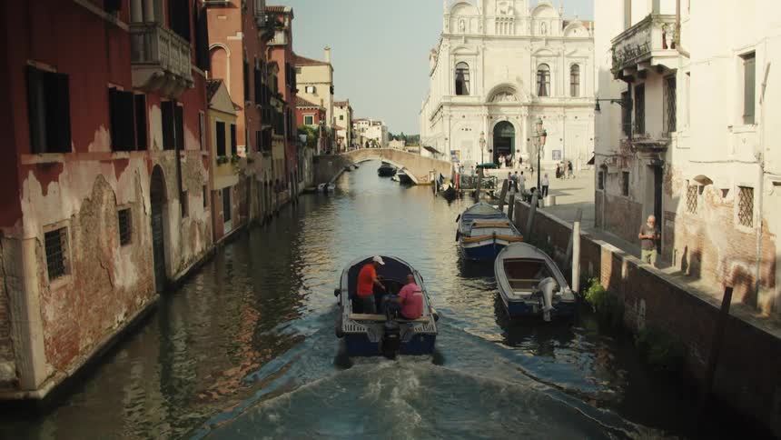Three men ride a motorboat through a narrow canal lined with old buildings