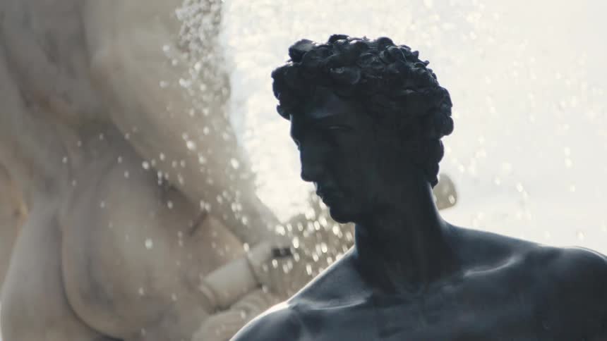 Backlit fountain spray sparkles behind a dark bronze male statue with curly hair and a pensive downward gaze