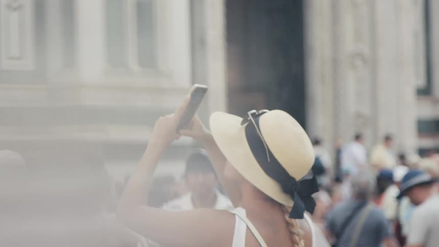 A person in a straw hat with a black ribbon lifts a phone to photograph an ornate cathedral entrance amid a busy crowd