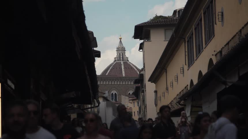 A crowded narrow street frames brunelleschis dome of santa maria del fiore in the distance the lantern gleaming above bustling shoppers beneath dark eaves