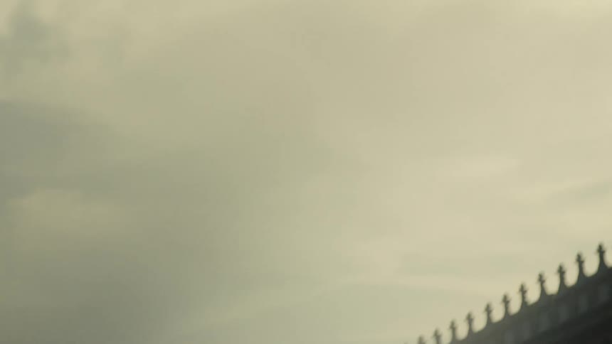 The video is an atmospheric blurry shot of the roofline of a building showing a row of silhouetted statues or architectural pinnacles against a hazy skybright dramatic beams of light cut through the thick fog or mist moving from the upper l